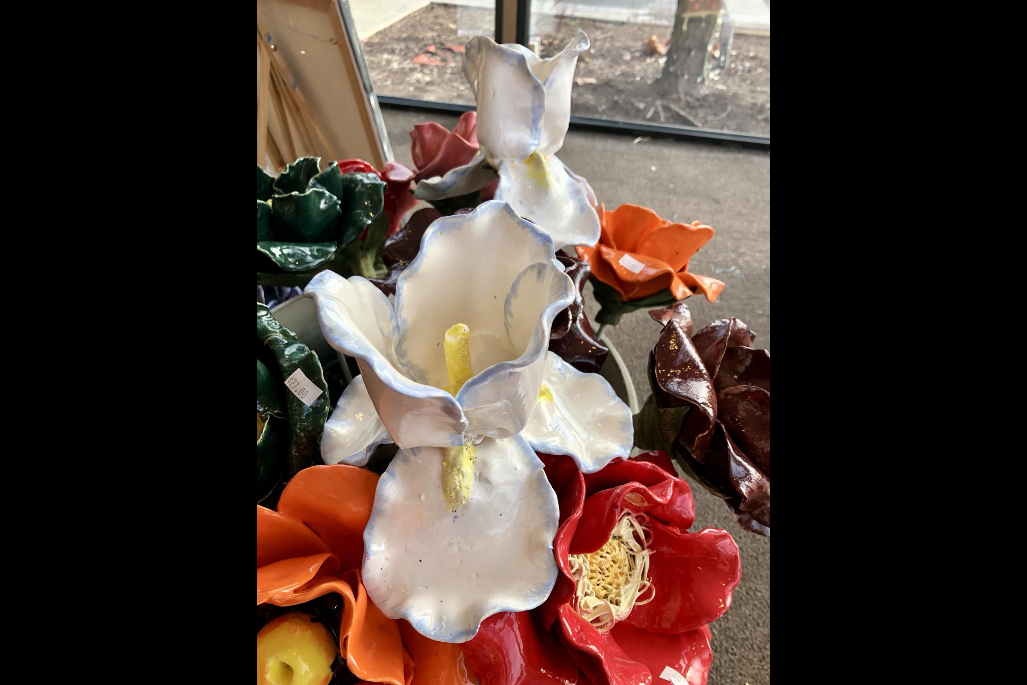 "A collection of handcrafted ceramic flowers displayed on a table near a window. The centerpiece features two large, white calla lily-inspired sculptures with delicate blue edges and yellow centers. Surrounding them are vibrant ceramic flowers in shades of red, orange, green, and brown, each intricately designed with unique textures and details. Small price tags are visible on some pieces. Natural light from the window highlights the glossy finish of the ceramics."