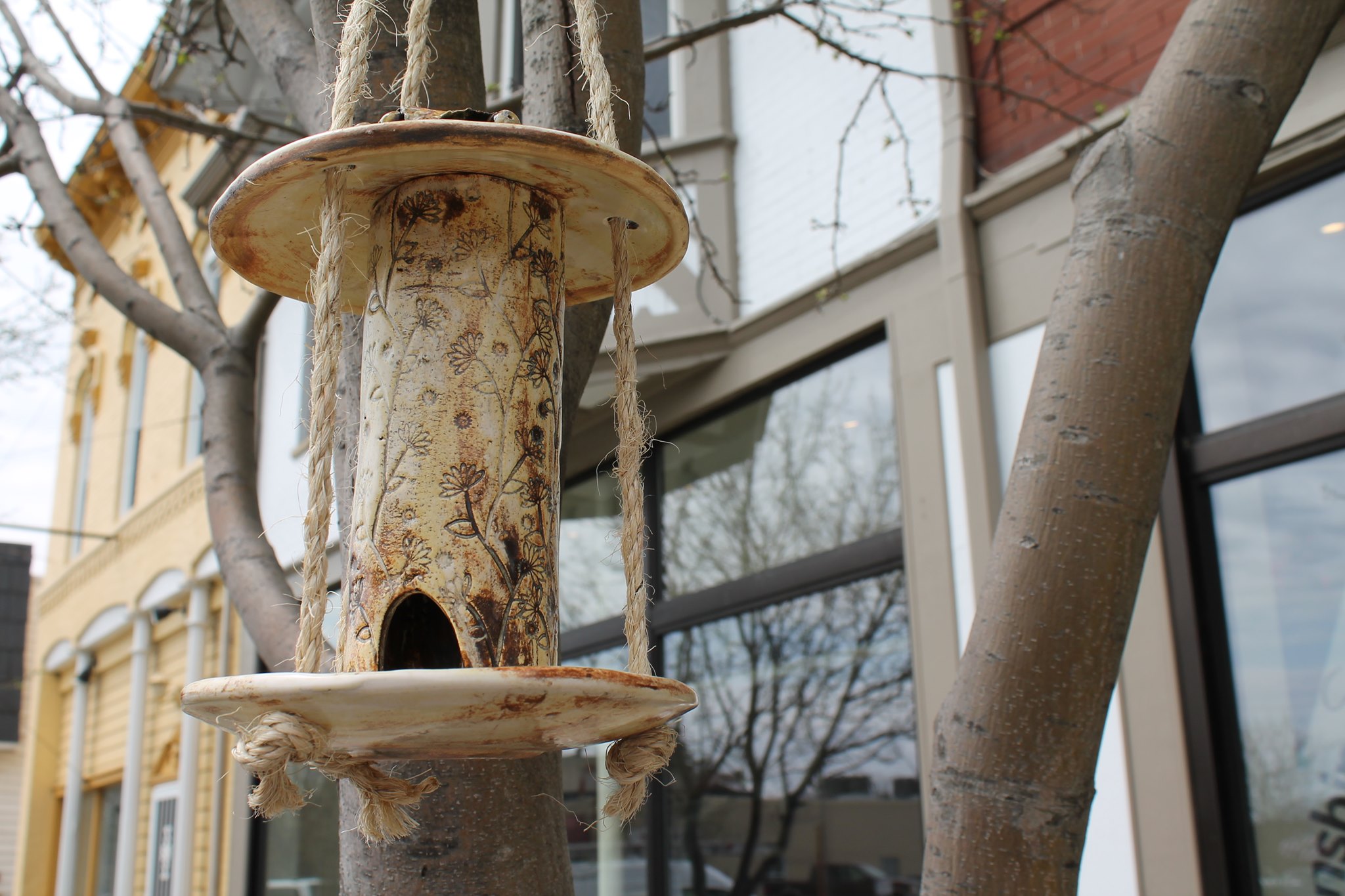 "Handcrafted clay bird feeder hanging from a tree branch in front of a building with large glass windows."