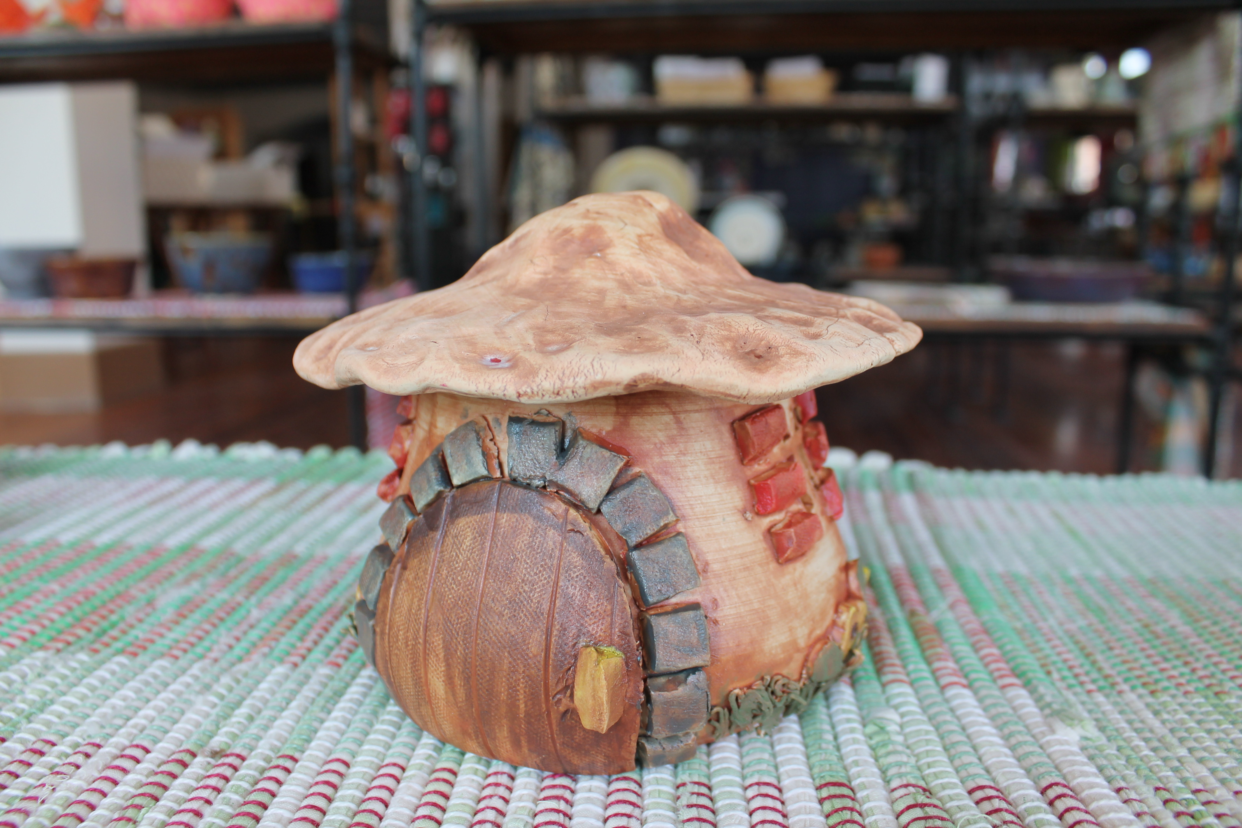 A handcrafted ceramic fairy house with a rustic wooden door, stone accents, and a mushroom-shaped roof, sitting on a colorful woven mat in a studio setting