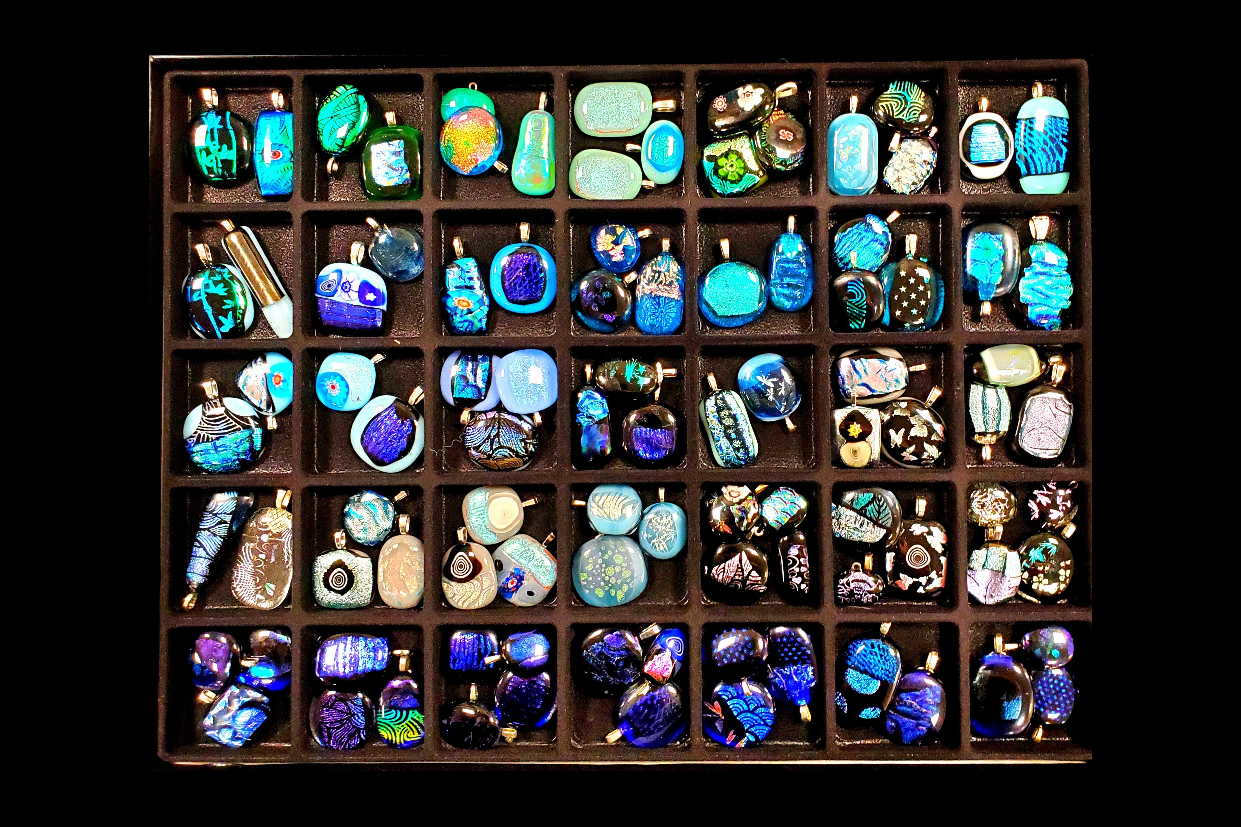 "Colorful fused glass jewelry pieces in shades of blue and green, neatly arranged in a display case."