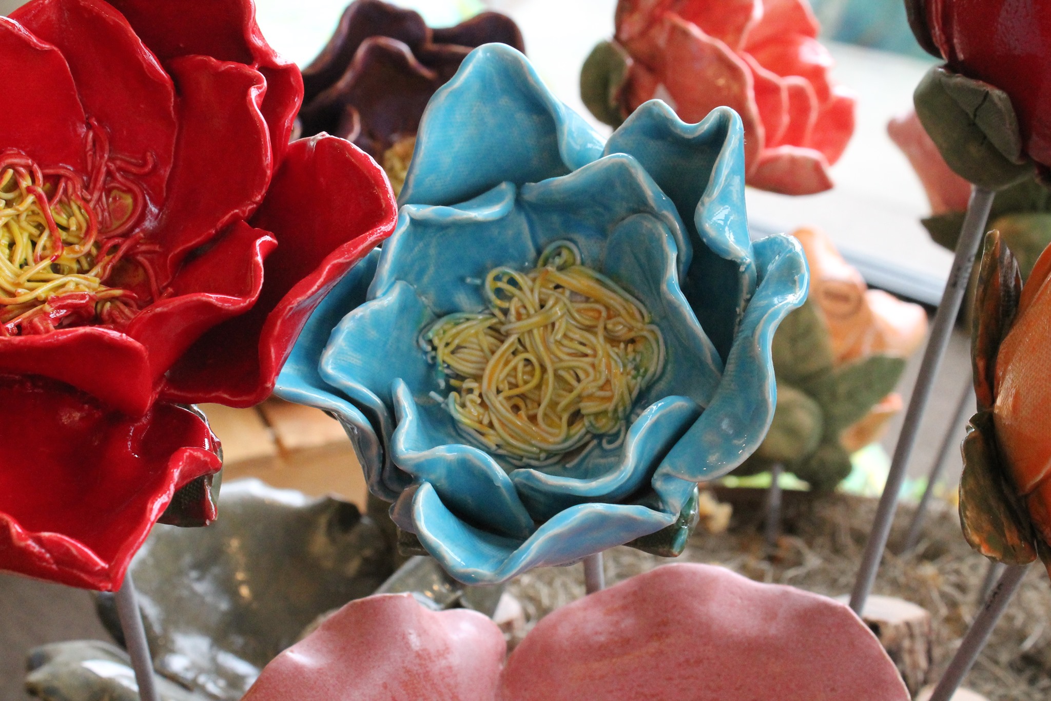 A close-up of vibrant ceramic garden flowers, including a bright red bloom and a blue flower with intricate detailing at the center. The petals are glossy and lifelike, showcasing the creativity and craftsmanship involved in their creation.