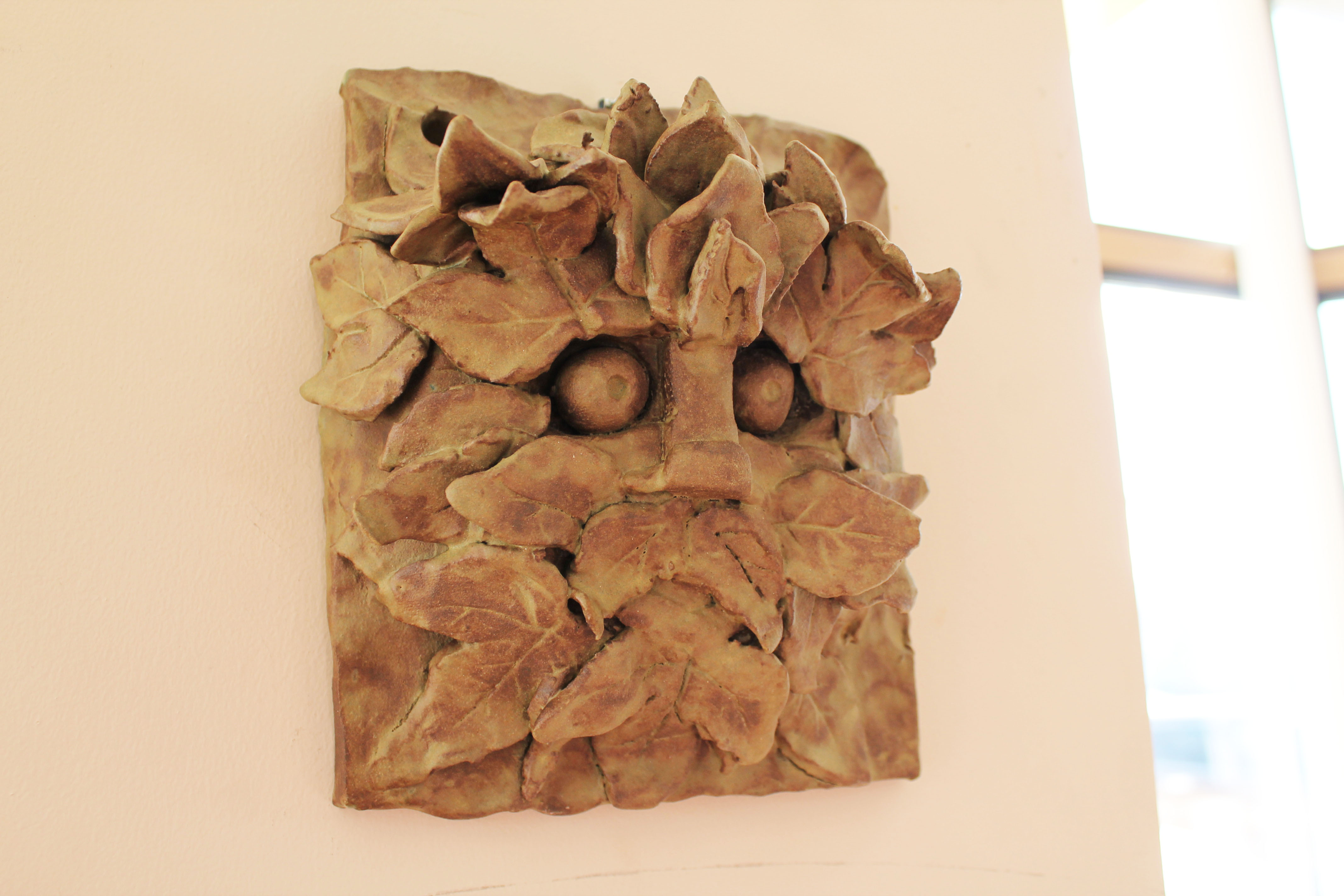 Handcrafted ceramic Greenman mask with intricate leaf detailing and earthy tones, mounted on a light wall.