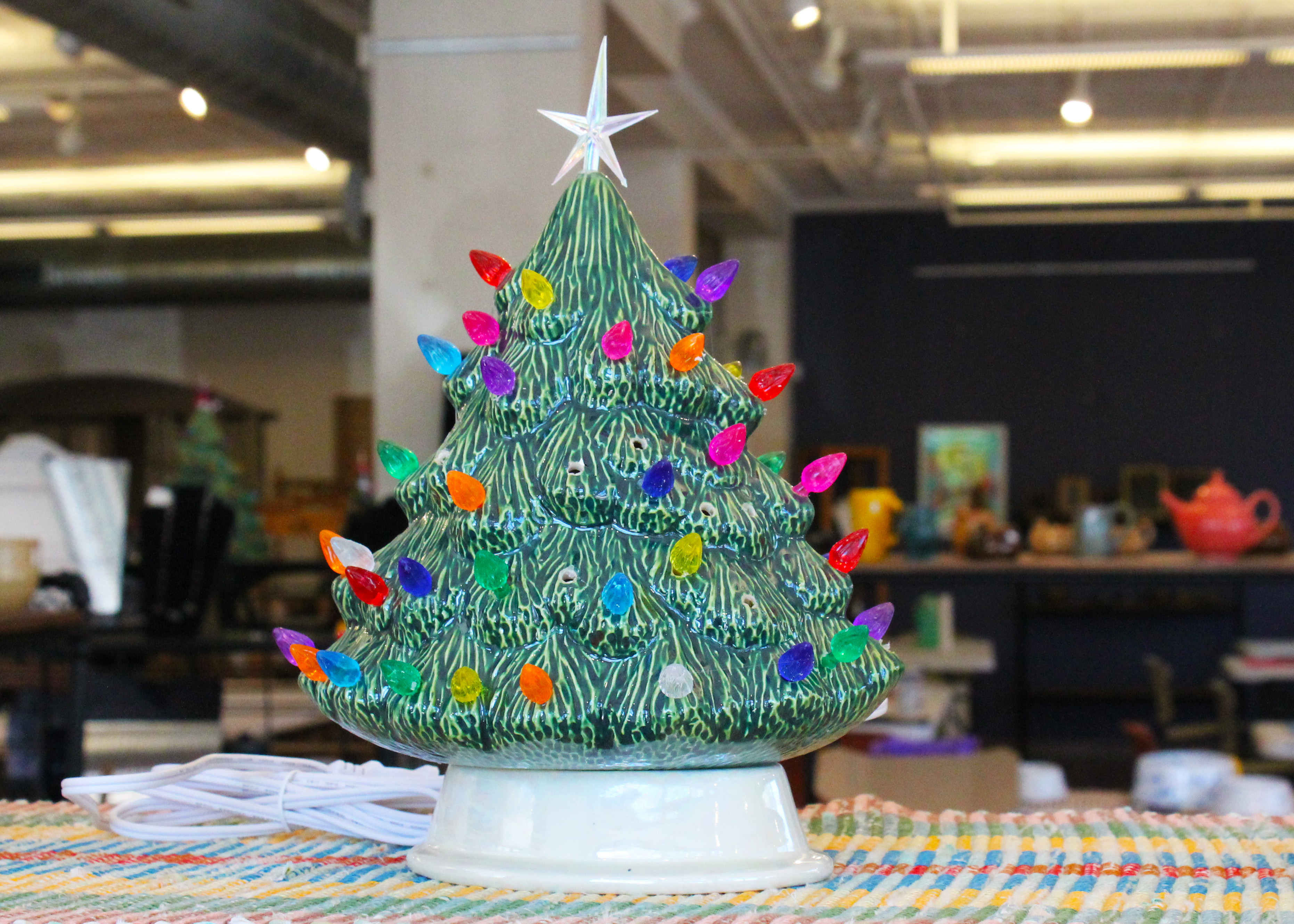 Alt Text: A painted ceramic Christmas tree adorned with colorful light bulbs and topped with a clear star. The tree sits on a white base in a bright, spacious art studio.