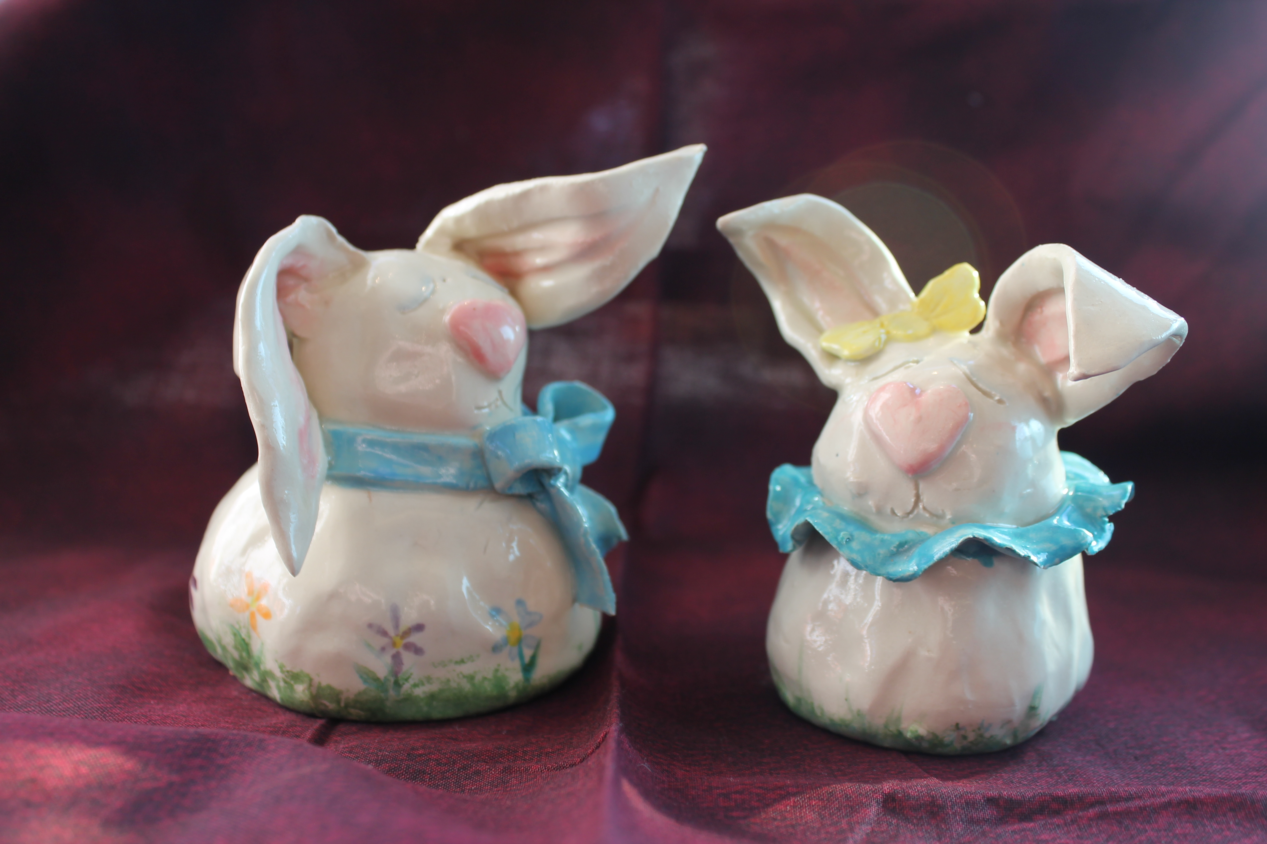 Two handcrafted ceramic pinch pot bunnies with painted floral accents, one wearing a blue bow and the other with a yellow bow, displayed on a rich burgundy background.