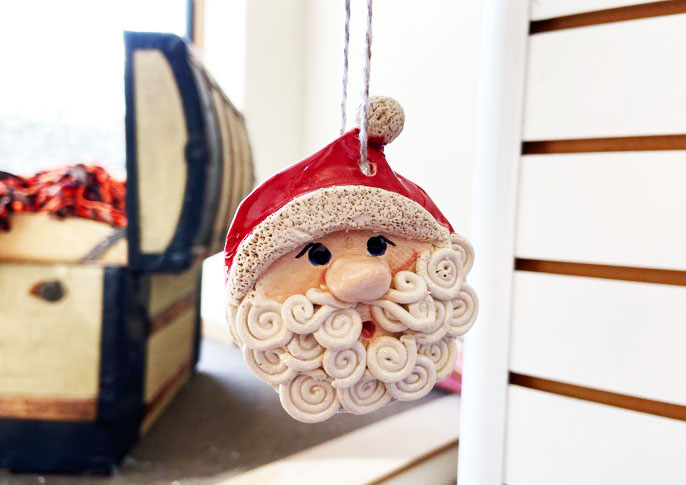 A handcrafted ceramic Santa Claus head ornament with a curly white beard, rosy cheeks, and a red hat, hanging by a string in a cozy, festive setting.