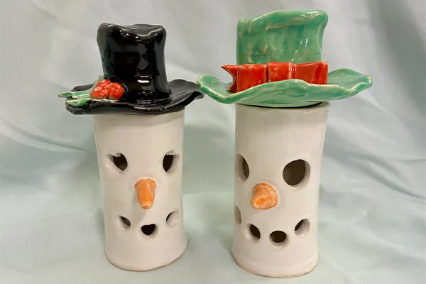 Two ceramic snowman luminaries with cylindrical white bodies featuring cut-out holes for eyes and mouths and small orange carrot noses.