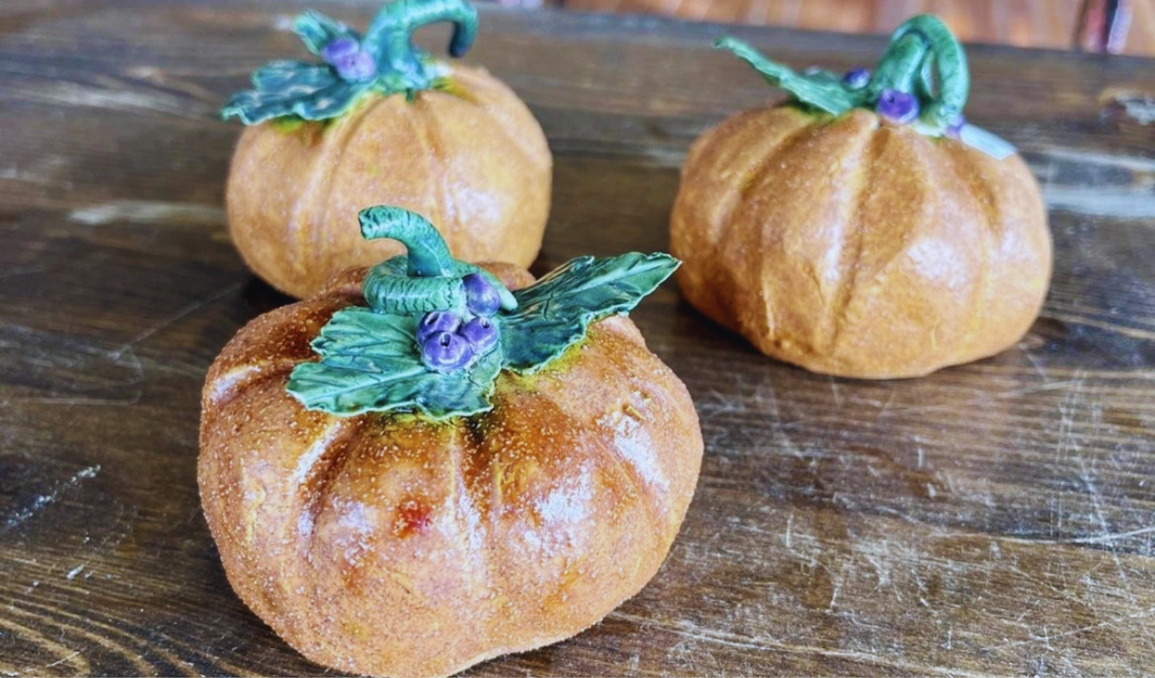 Three handmade ceramic pinch pot pumpkins with green leaves and purple accents sit on a wooden table, showcasing finished pieces from a Sunshine Studios fall pottery class.