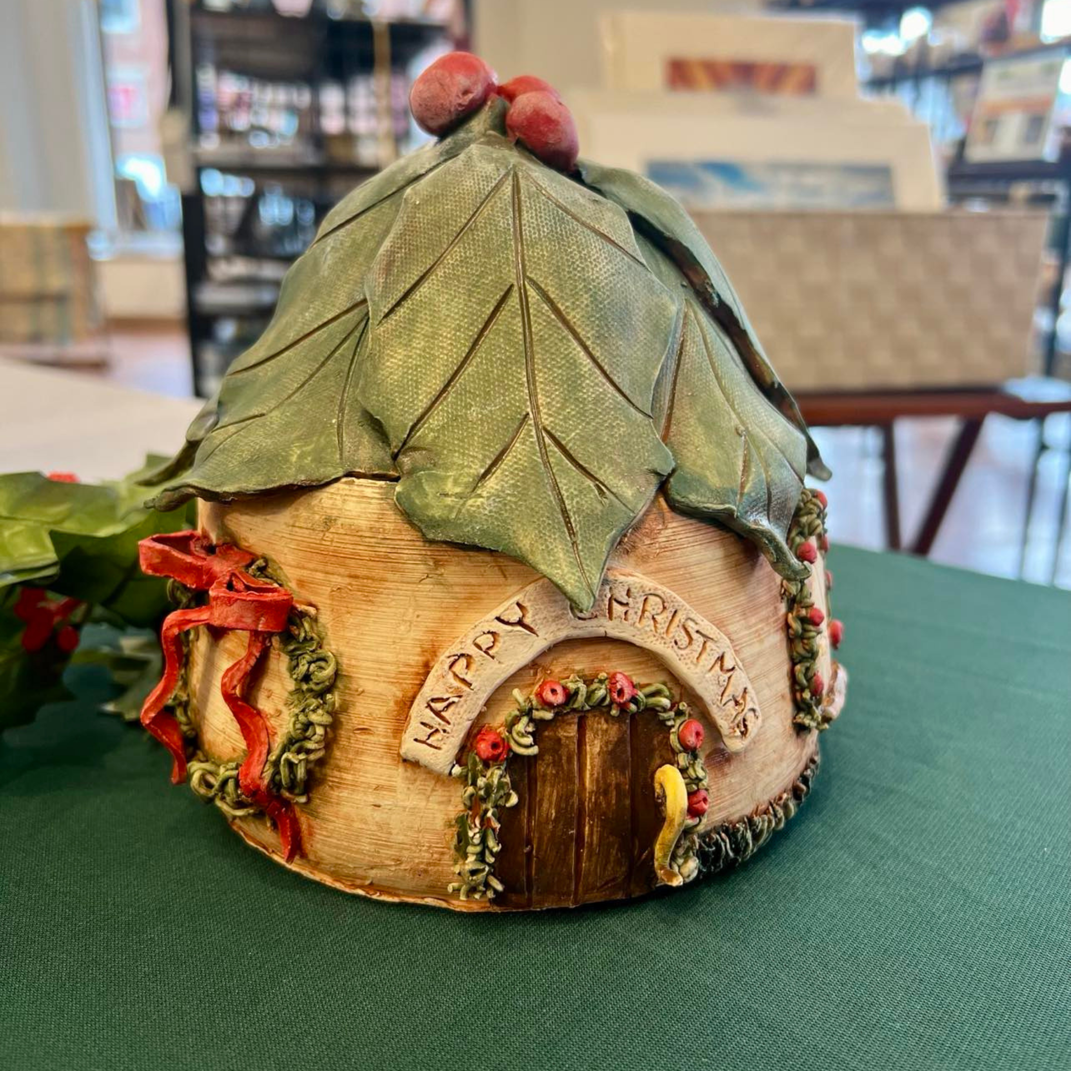 A miniature fairy house decorated for Christmas, featuring a leaf-textured green roof with two red berries on top.