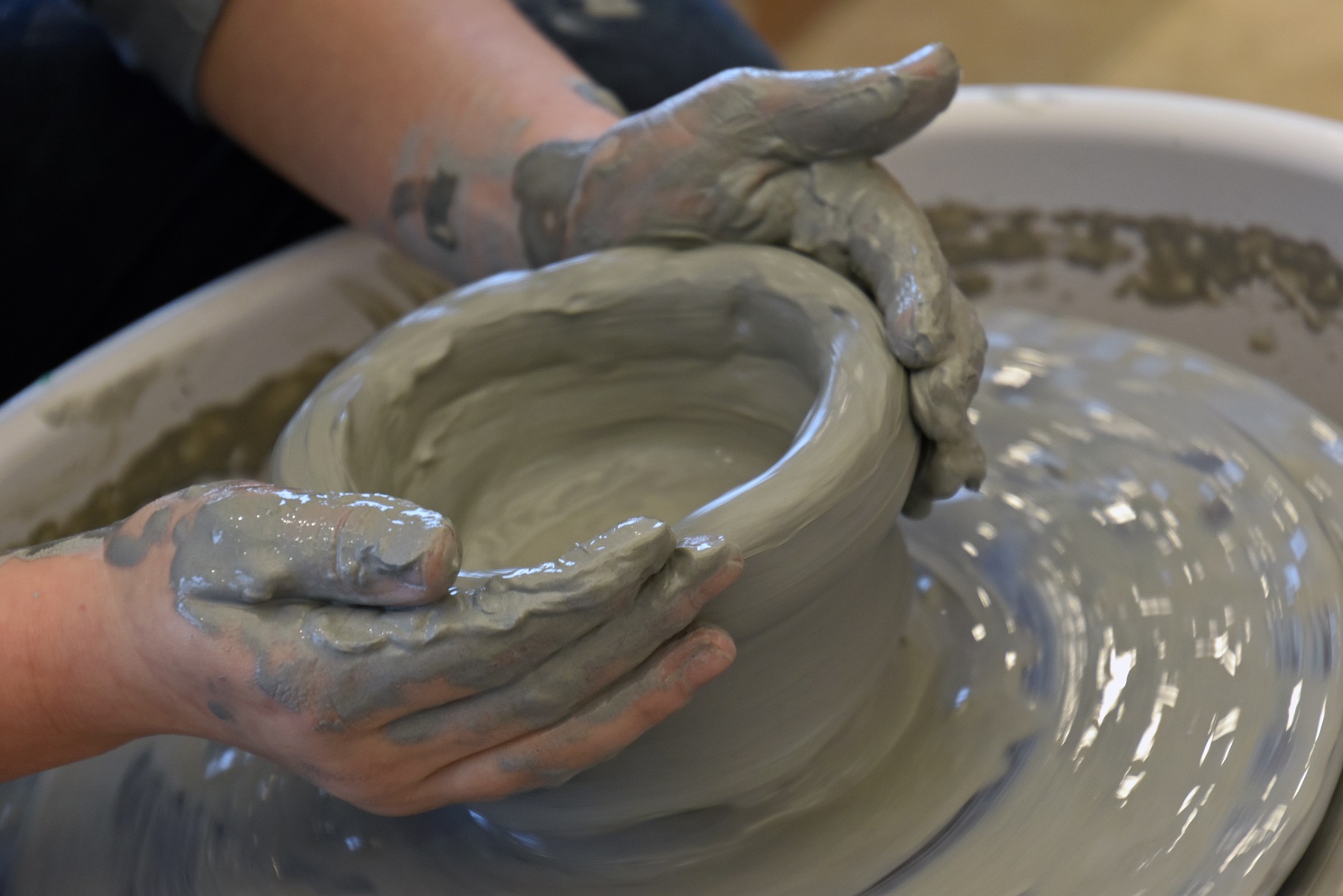 Hands shaping a clay pot on a pottery wheel, with wet clay in motion, showcasing the art of wheel throwing.