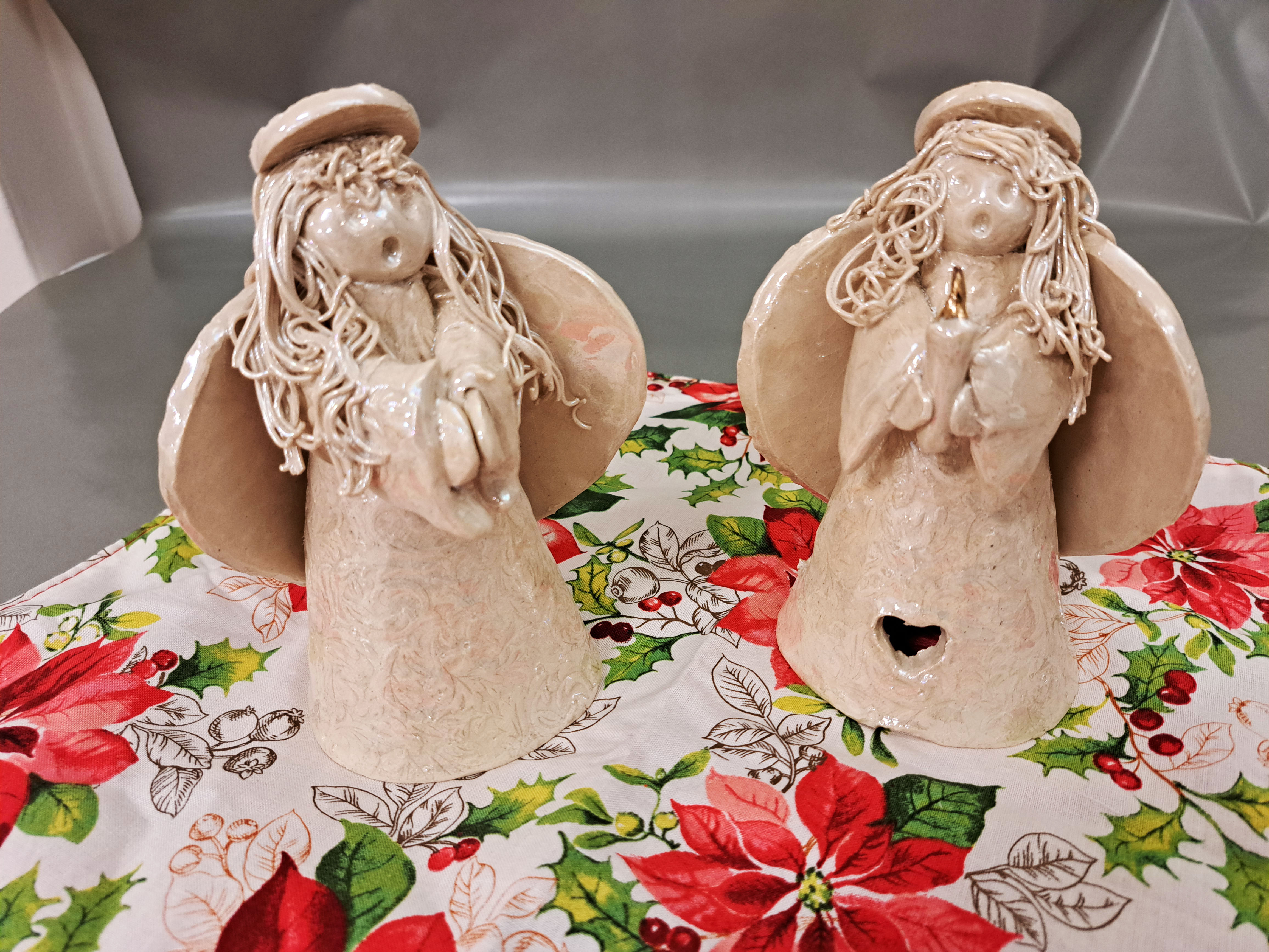 Two handcrafted ceramic angel figurines with intricate details, each holding a small candle. The angels have textured wings, flowing hair, and serene expressions, placed on a festive cloth decorated with poinsettias and holly.