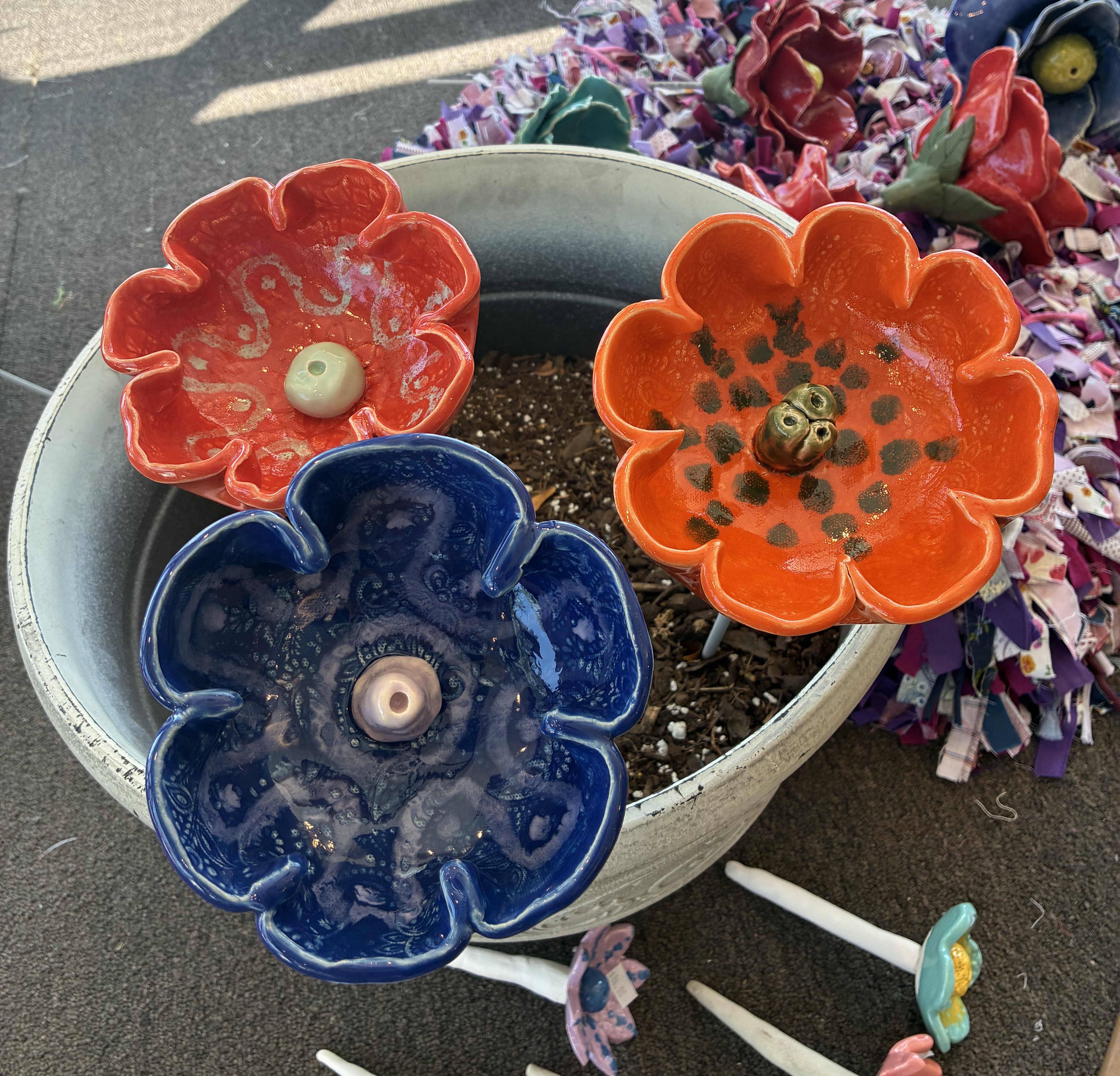 Three colorful ceramic flower-shaped bird baths in orange, coral, and blue with decorative glazed centers, displayed on garden stakes.