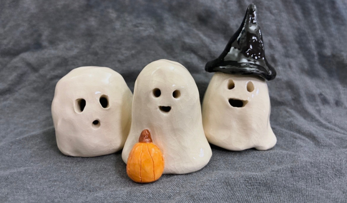 Three handmade ceramic ghost figures with playful facial expressions, including one wearing a witch hat, displayed with a small pumpkin as festive Halloween pottery.