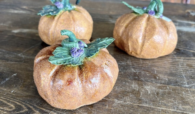 Three handmade ceramic pinch pot pumpkins with sculpted leaves and curled stems, displayed on a wooden table as decorative fall pottery.
