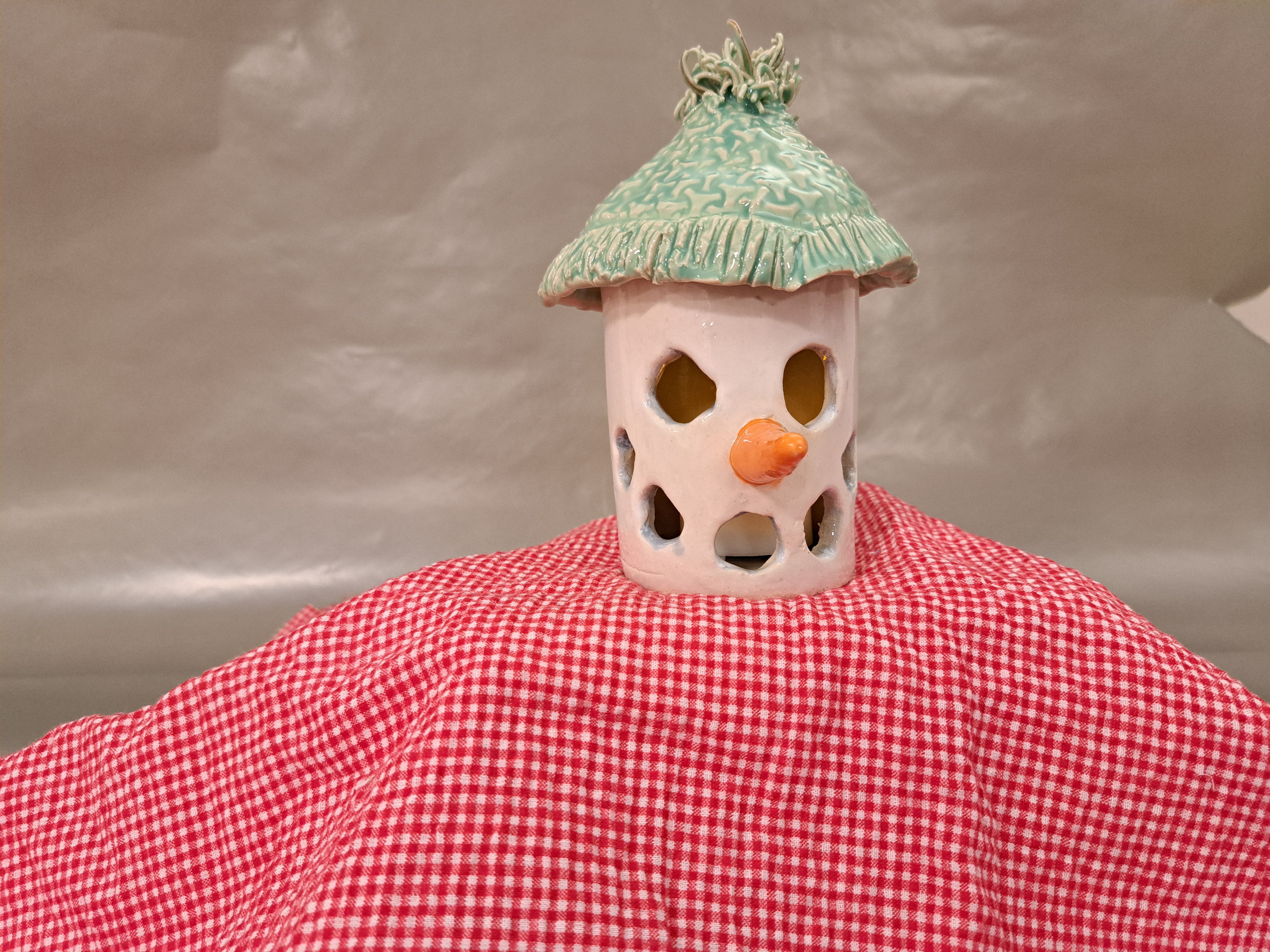 Alt text: A handcrafted ceramic snowman luminary with a carrot-shaped nose and cutout holes for light, topped with a textured green hat, displayed on a red gingham cloth.