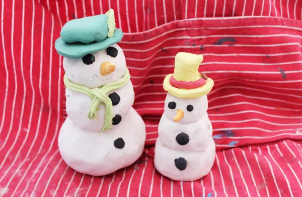 Two handcrafted clay snowmen, one large and one small, both with black coal buttons and eyes, orange carrot noses, and brightly colored hats. The larger snowman wears a green hat and scarf, while the smaller snowman has a yellow hat with a red band. They are set against a red and white striped fabric background