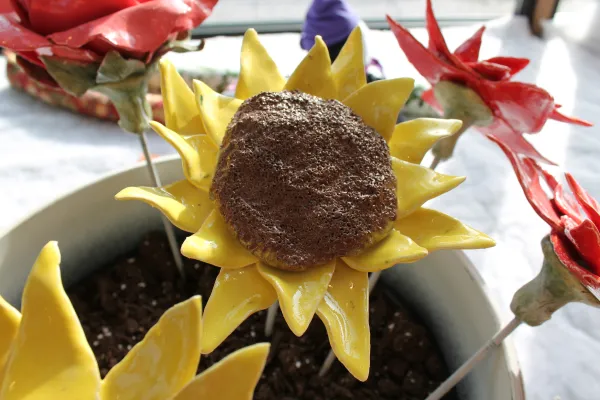 A vibrant clay sunflower with glossy yellow petals and a textured brown center is displayed in a pot, surrounded by other colorful clay flowers. The intricate details showcase the craftsmanship and creativity involved in making the piece.