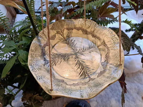 A handcrafted ceramic hanging bird bath with an earthy beige glaze, suspended by twine strings. The surface features intricate imprints of pine needles at the center and a textured border with tree-like patterns. The bird bath hangs amid lush green foliage, with a snowy outdoor scene visible in the background."