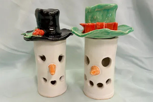 Two ceramic snowman luminaries with cylindrical white bodies featuring cut-out holes for eyes and mouths and small orange carrot noses.