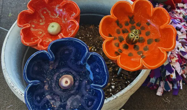 Three handmade ceramic flower-shaped bee baths in bright orange and deep blue glazes are displayed in a planter, each designed to provide a safe drinking spot for bees and other pollinators