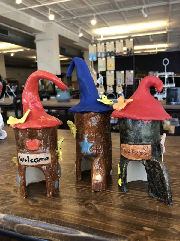 Three whimsical ceramic fairy houses shaped like tree trunks with colorful curved roofs, tiny doors, butterflies, and welcome signs displayed on a table.