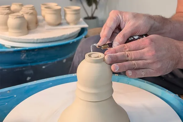 Close-up of a potter's hands covered in wet clay, shaping a vessel on a spinning pottery wheel. The soft clay is forming into a rounded pot as the wheel rotates, capturing the hands-on process of wheel throwing in ceramics.
