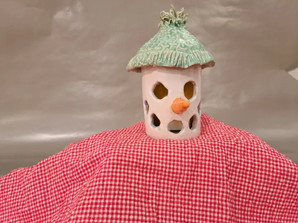 Alt Text: A handcrafted ceramic snowman luminary with a green textured hat, a carrot-shaped nose, and circular cutouts for light. It sits atop a red and white checkered fabric.