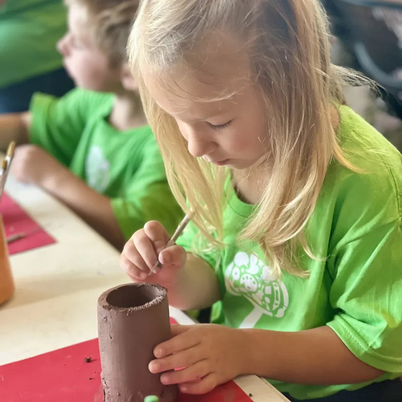 A young child wearing a bright green shirt carefully paints a clay cylinder during an art activity.