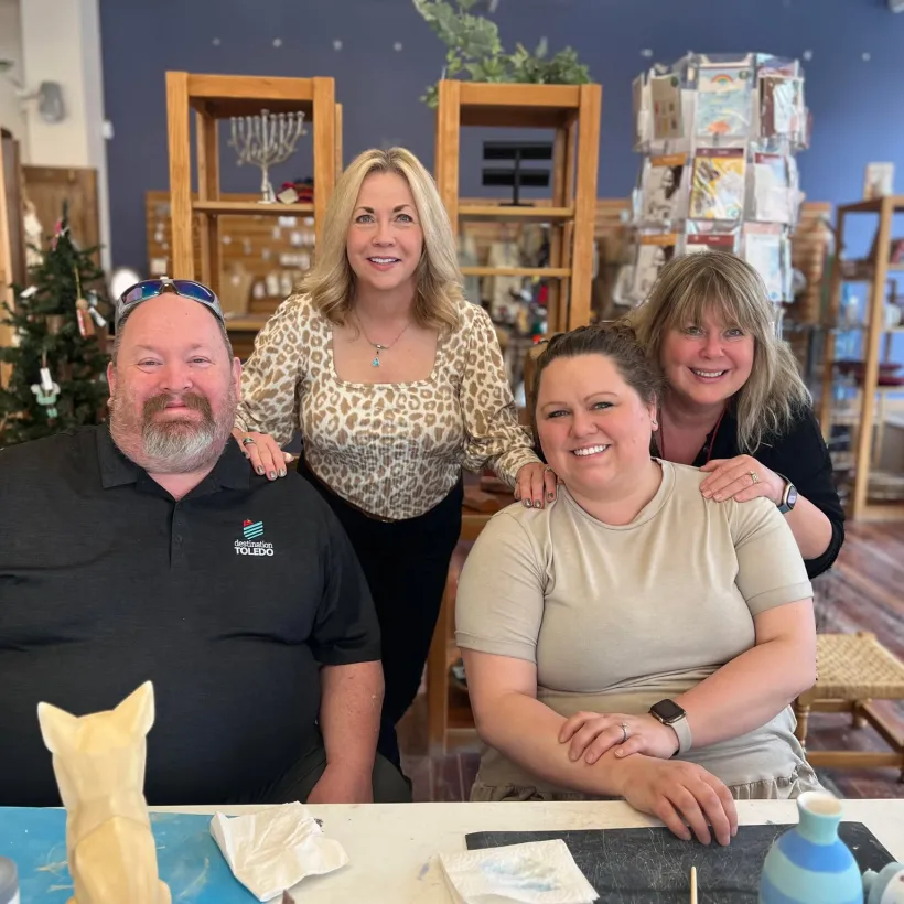 Four adults smile together inside a craft or pottery studio. Two are seated at a table with paint and ceramic pieces, while two stand behind them with their hands on the seated individuals' shoulders.