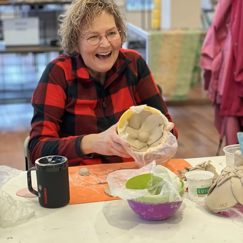 A person wearing a red and black plaid shirt smiles while working on a clay project at a table.