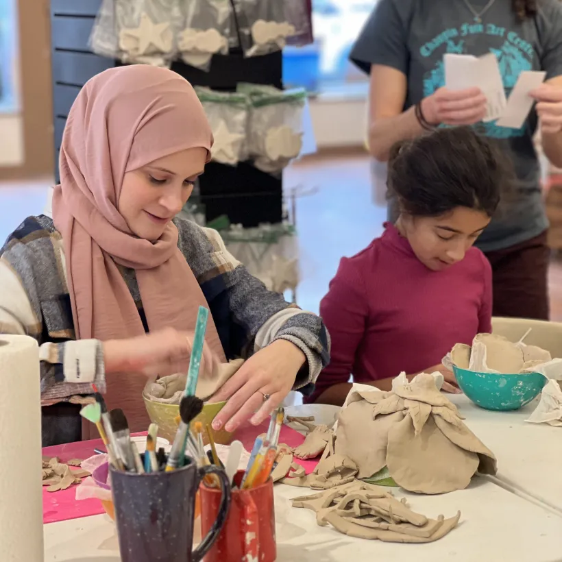 A woman and a young girl work with clay at a table covered in pottery tools and brushes. The woman smiles as she shapes clay, while the girl focuses on her own project.